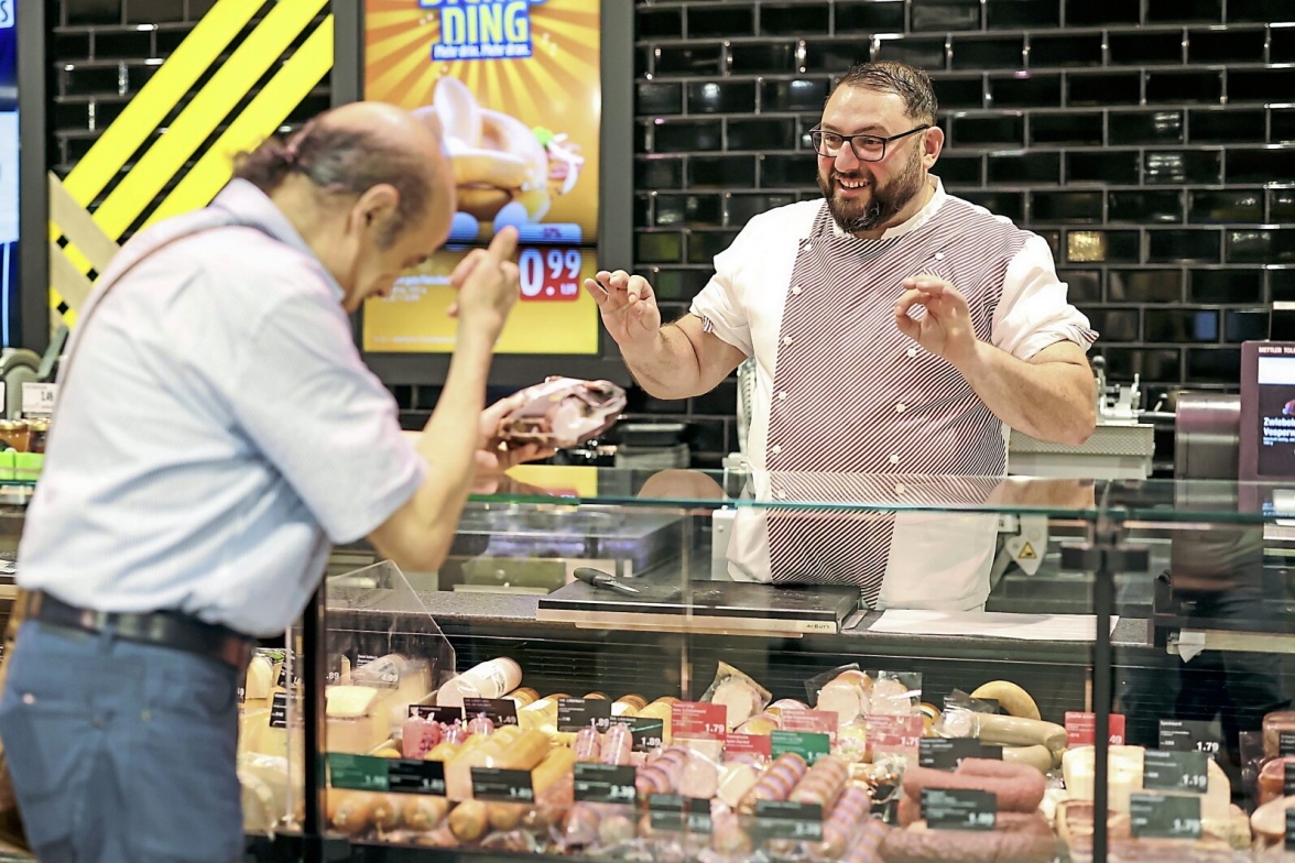Metzger Yevgeniy Klibanov (r) lernt Gebärdensprache, um sich an der Theke im Supermarkt mit gehörlosen Kunden wie Stammkunde Hedi Doudeche (1) zu unterhalten. Foto: Daniel Löb/dpa/dpa-Bildfunk
