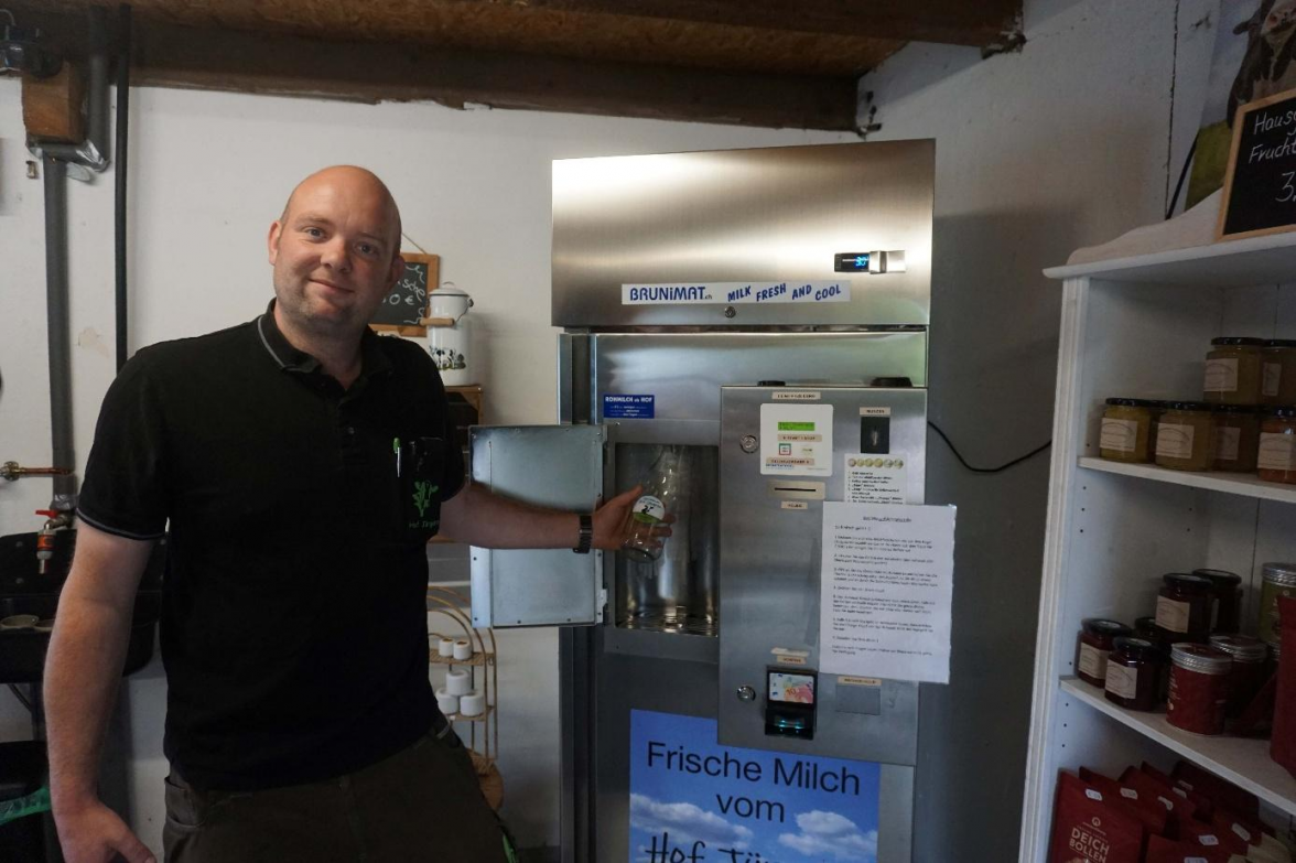 Thorben Jürgens präsentiert die hofeigene Milchtankstelle, wo sich Genießerinnen und Genießer die täglich frisch eingefüllte Rohmilch selbst abzapfen können. Bild: Friedhelm Müller-Düring