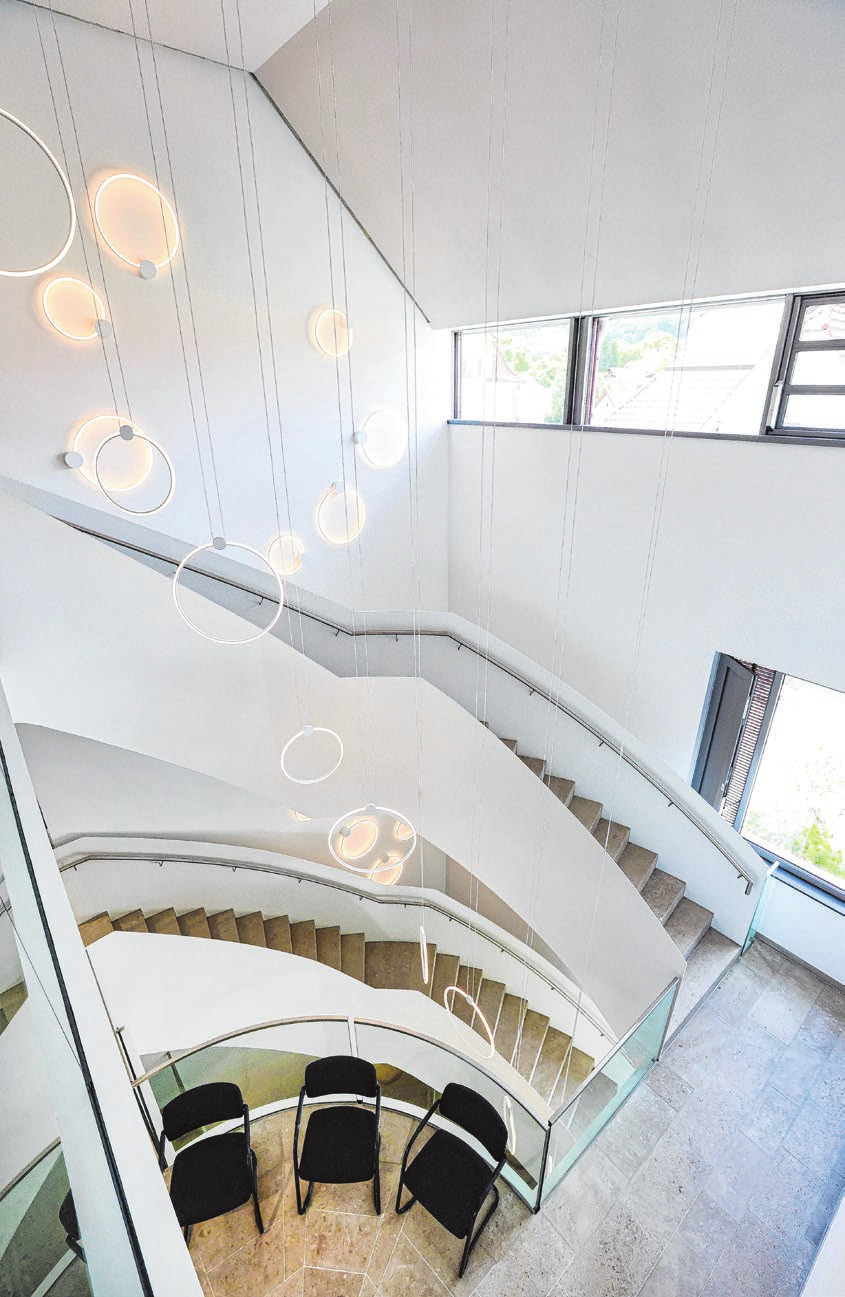 Eine geschwungene Treppe verbindet elegant alle Stockwerke im Neubau. Ringförmige Leuchten setzen den architektonischen Blickfang zudem gekonnt in Szene.