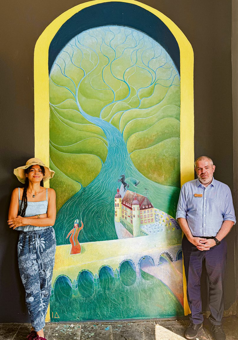 Künstlerin Isabel Antonio und Hoteldirektor Benjamin Simmer vor dem neuen Mural „Confluentes“ am City Hotel Kurfürst Balduin. Das rund 3,5 mal 1,7 Meter große Wandbild wurde im Juni 2026 fertiggestellt. Foto: City Hotel Kurfürst Balduin