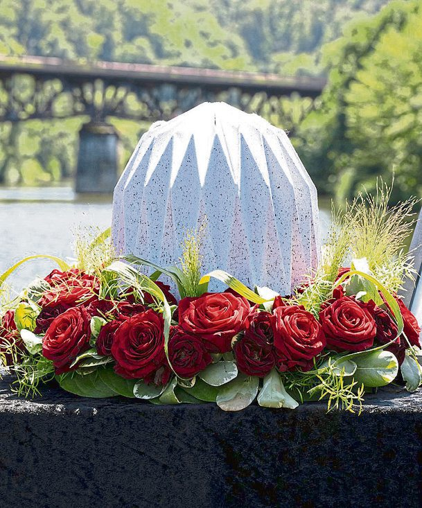 Eine von roten Rosen umgebene Urne am Fluss: Bei einer Flussbestattung wird die Asche in einer wasserlöslichen Urne dem Wasser übergeben. Die Trauerfeier kann auf einem Schiff stattfinden. Foto: Ruhewald Lahnhöhe Scheidt
